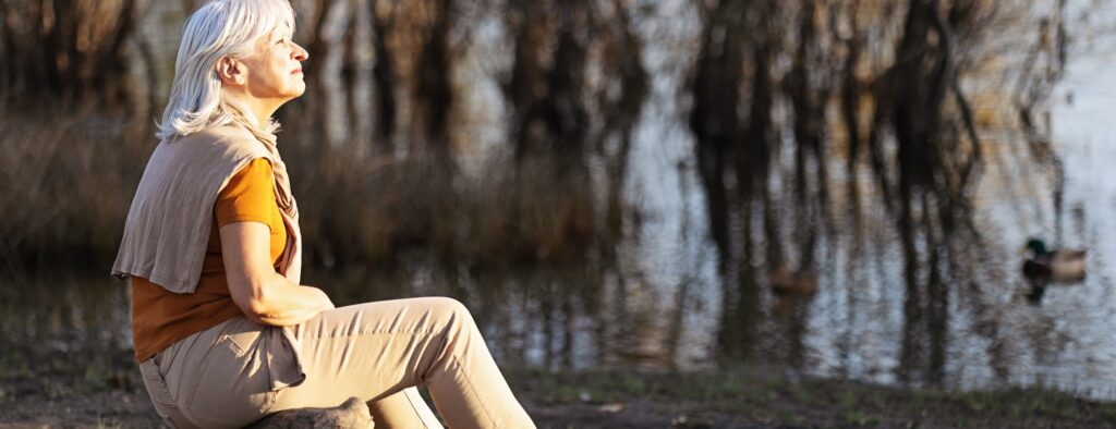 Woman over 50 sitting thoughtfully near a lake, reflecting on trust, surrender, and the next step in life.